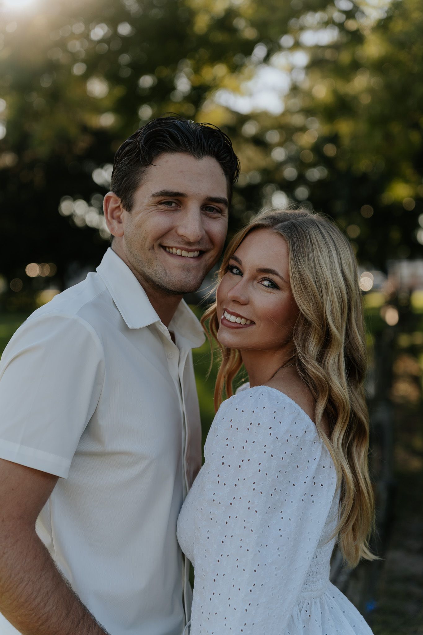 Smiling couple in white outfits posing closely together outdoors in golden hour sunlight.