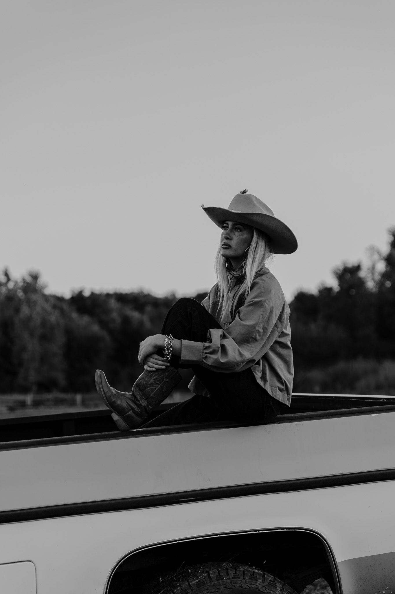 Black and white photo of woman in cowboy hat and boots sitting on a truck bed at dusk.