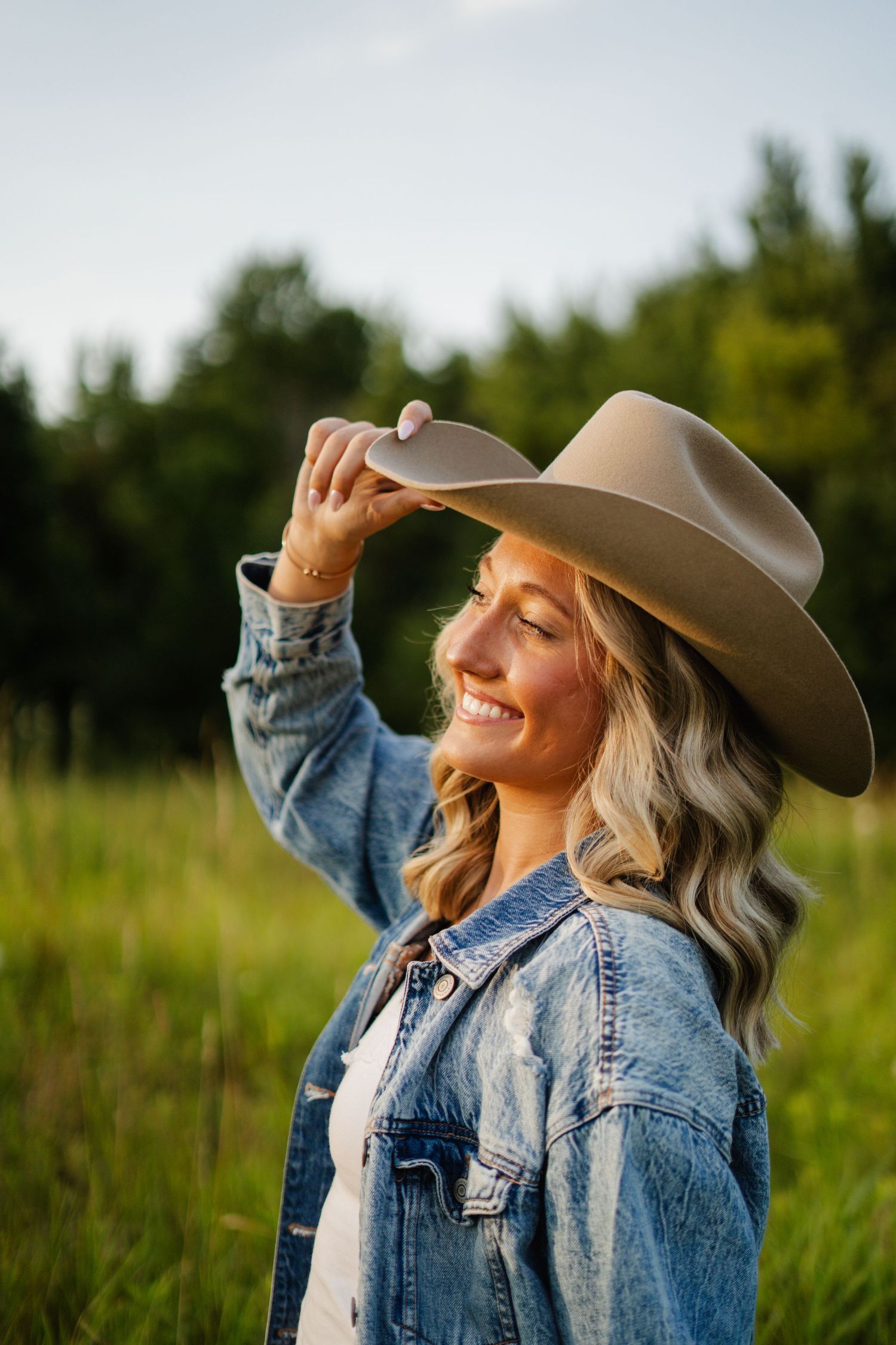 Joyful blonde woman tipping her tan cowboy hat while smiling in a sunlit green meadow.