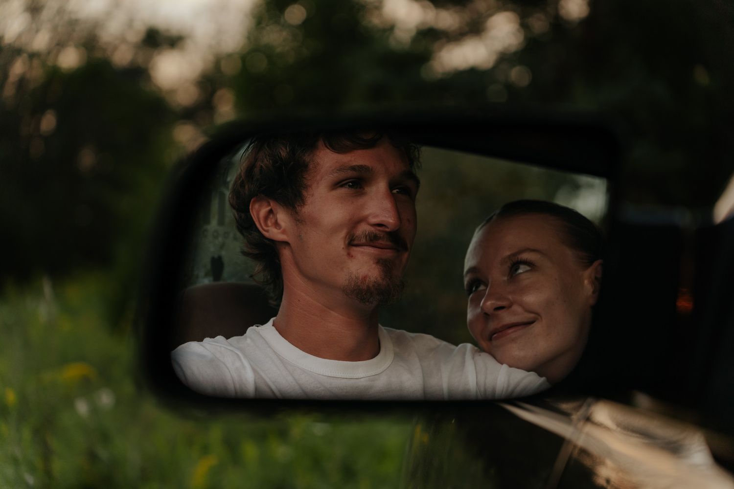 A happy couple reflected in a car side mirror, smiling at each other in a green setting.