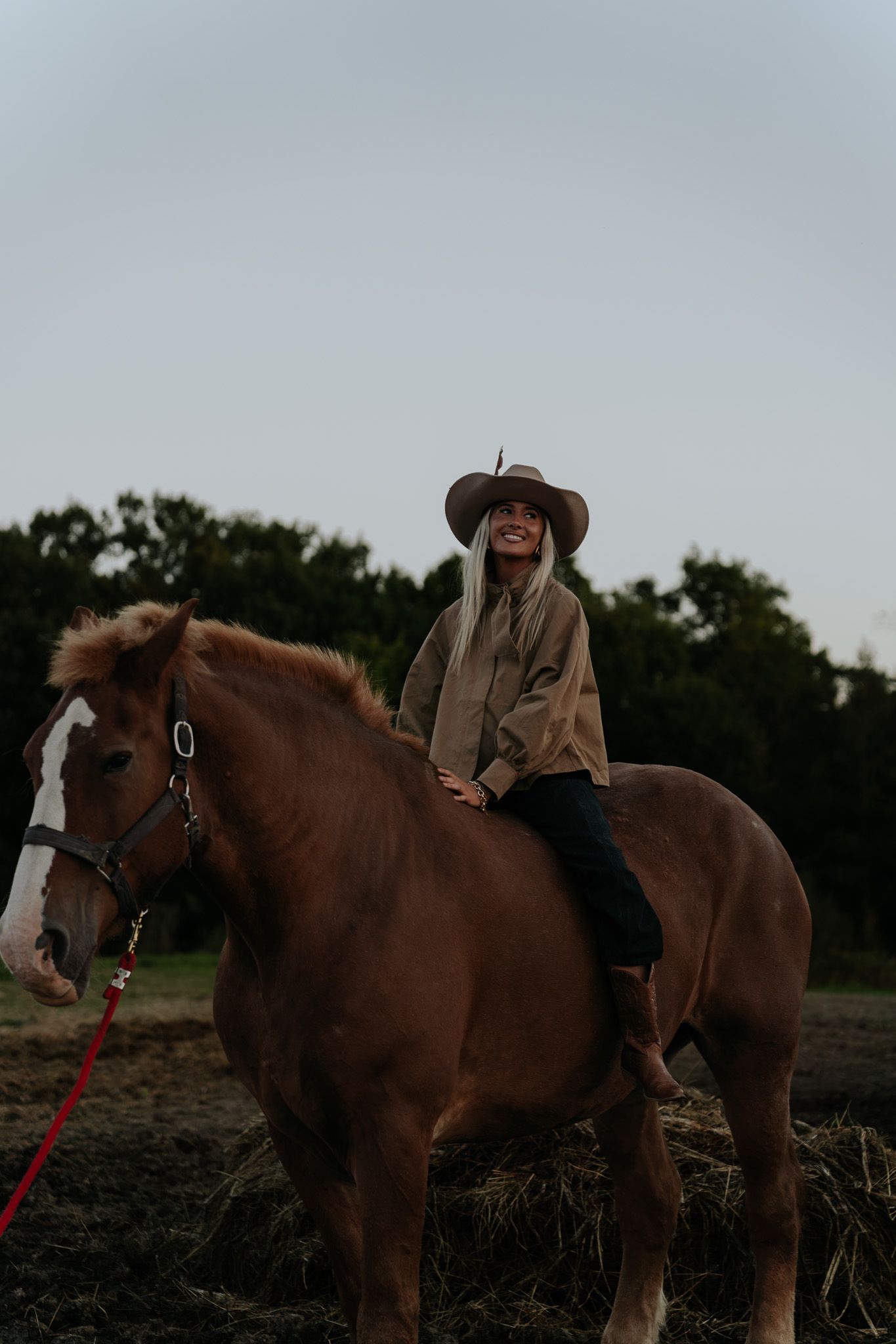 Smiling blonde woman in cowboy hat riding a chestnut horse with a red lead rope on a farm.
