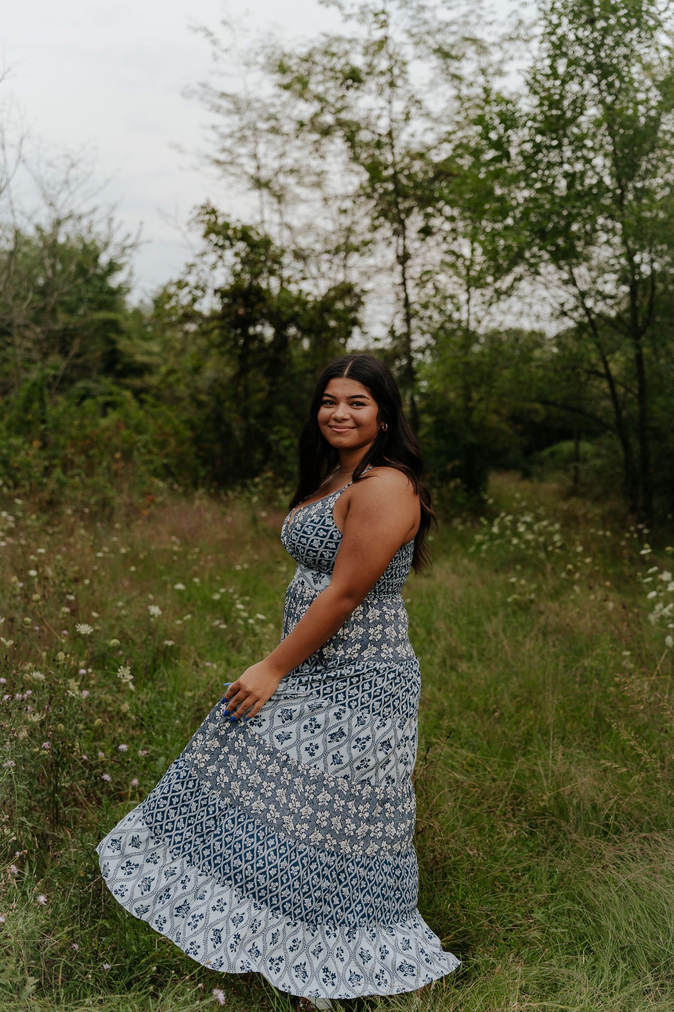 Smiling young woman in blue floral maxi dress twirling in lush green field for senior photos.