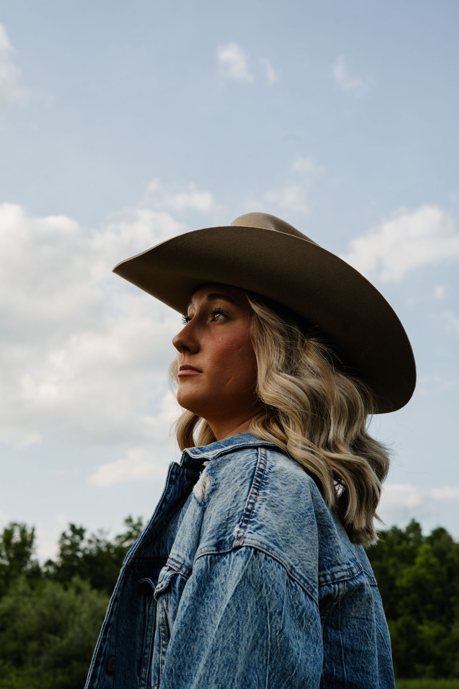 Blonde woman in brown cowboy hat and denim jacket gazing upward against cloudy sky outdoors.