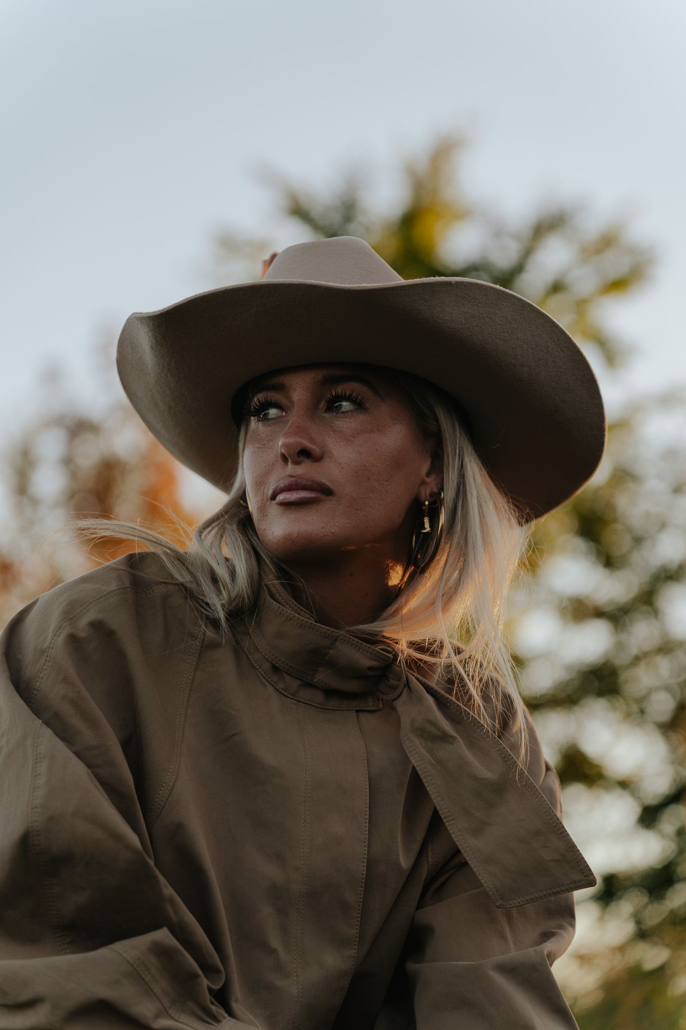Woman in wide-brim cowboy hat and olive trench coat gazing upward outdoors in autumn.