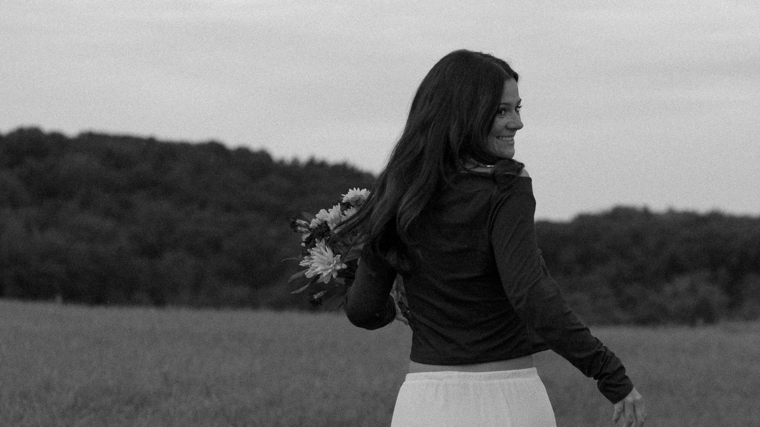 Black and white photo of a woman twirling in a field holding wildflowers with trees in background.