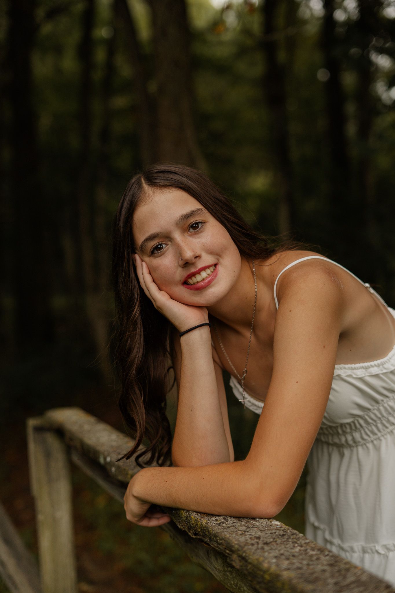 Smiling teen girl in white dress leaning on rustic wooden fence in forest.