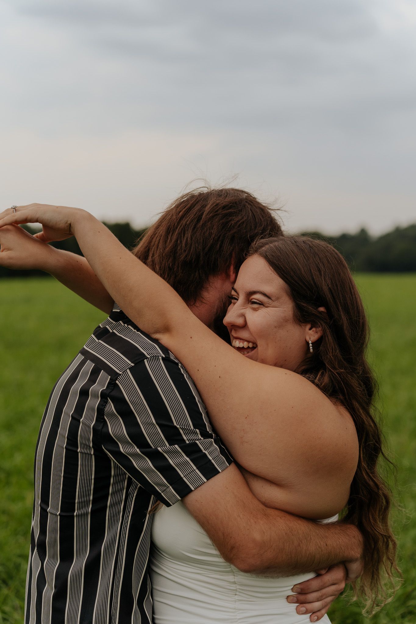 Laughing couple sharing a warm embrace in green field during golden hour outdoor engagement photography session.