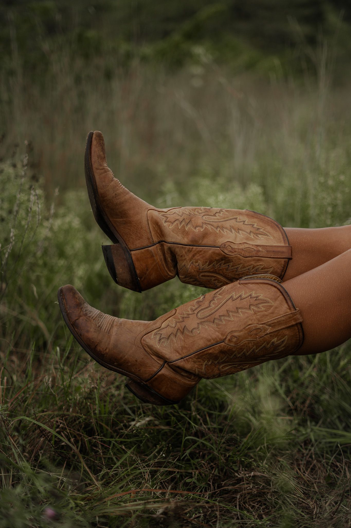 Close-up of tan cowboy boots with intricate stitching raised up against a lush green meadow.
