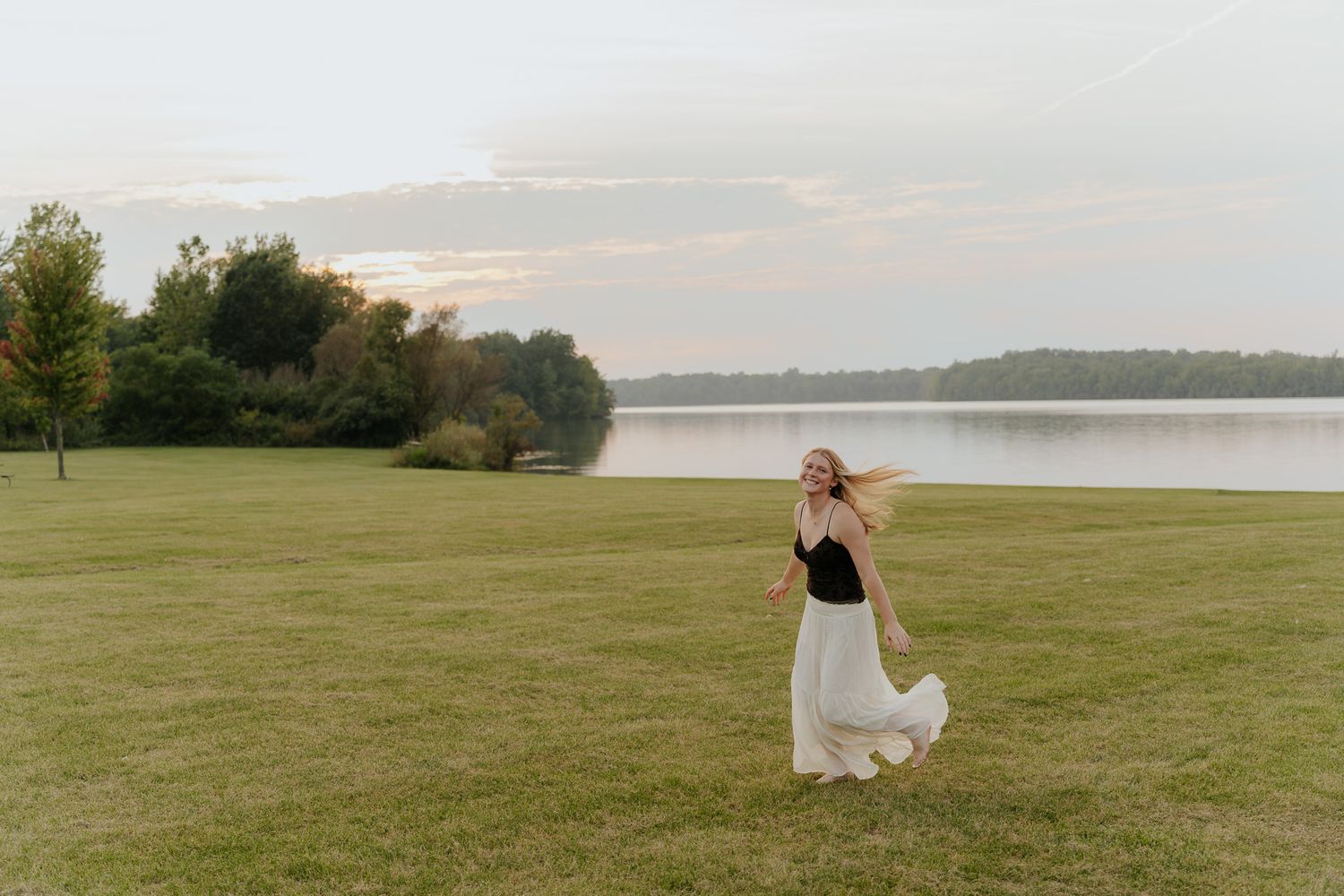 Blonde woman in black top and white maxi skirt twirling joyfully on green lawn beside a serene lake.