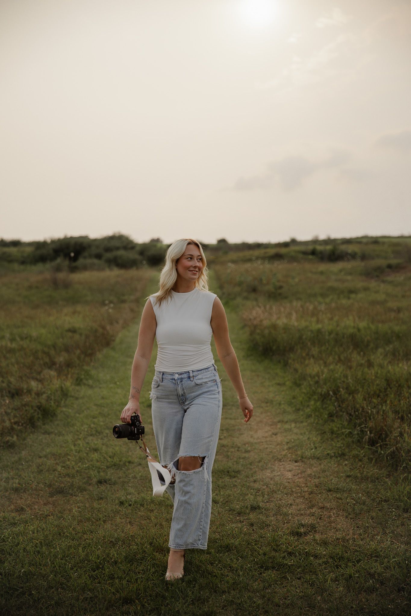 Smiling blonde woman walking through green field holding camera in ripped jeans and white top.