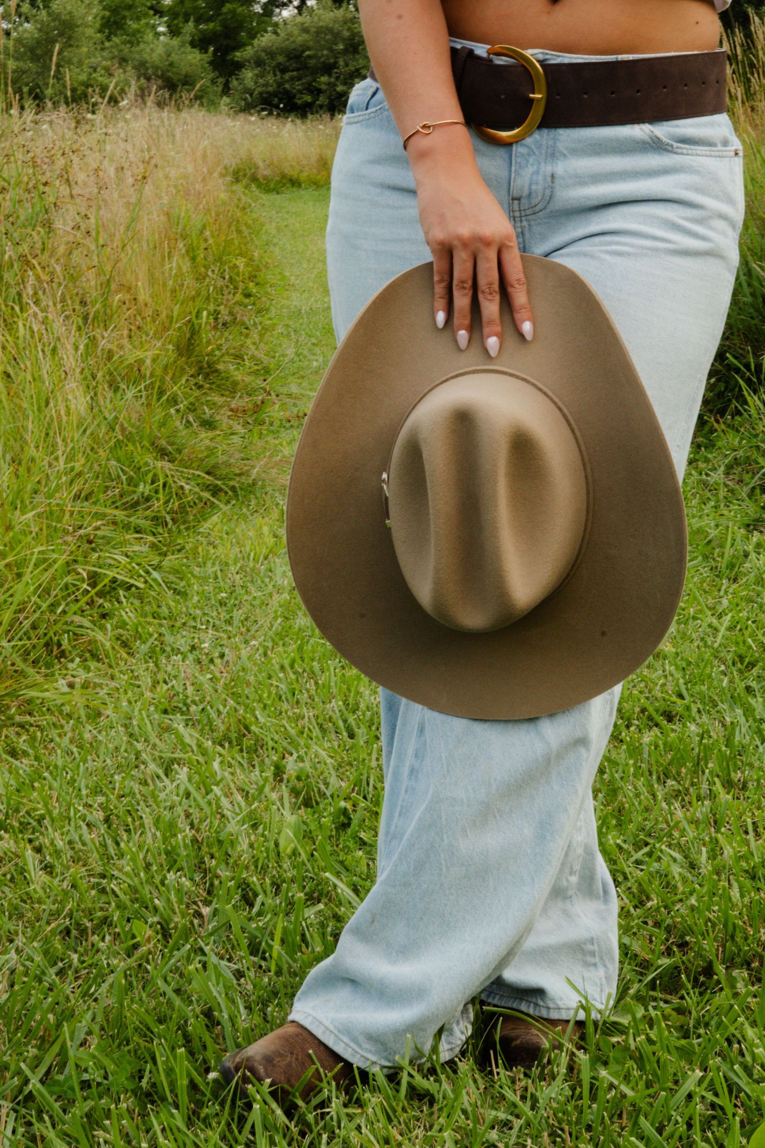 Close-up of woman in jeans and boots holding a tan cowboy hat in a grassy field.