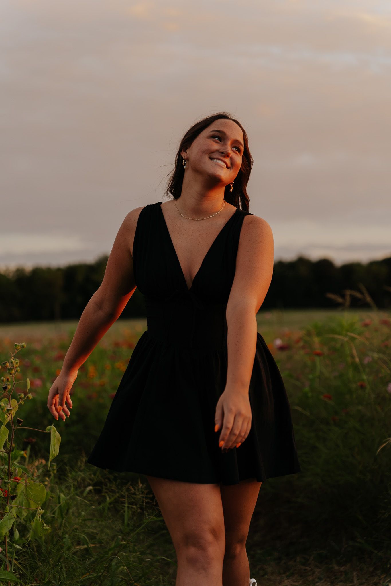 Young woman in black mini dress smiling in a field during a warm sunset with dramatic sky.