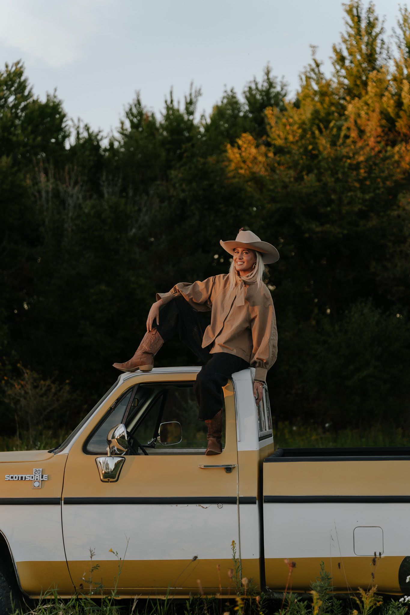 Smiling blonde woman in cowboy hat sitting atop a vintage yellow and white pickup truck outdoors.