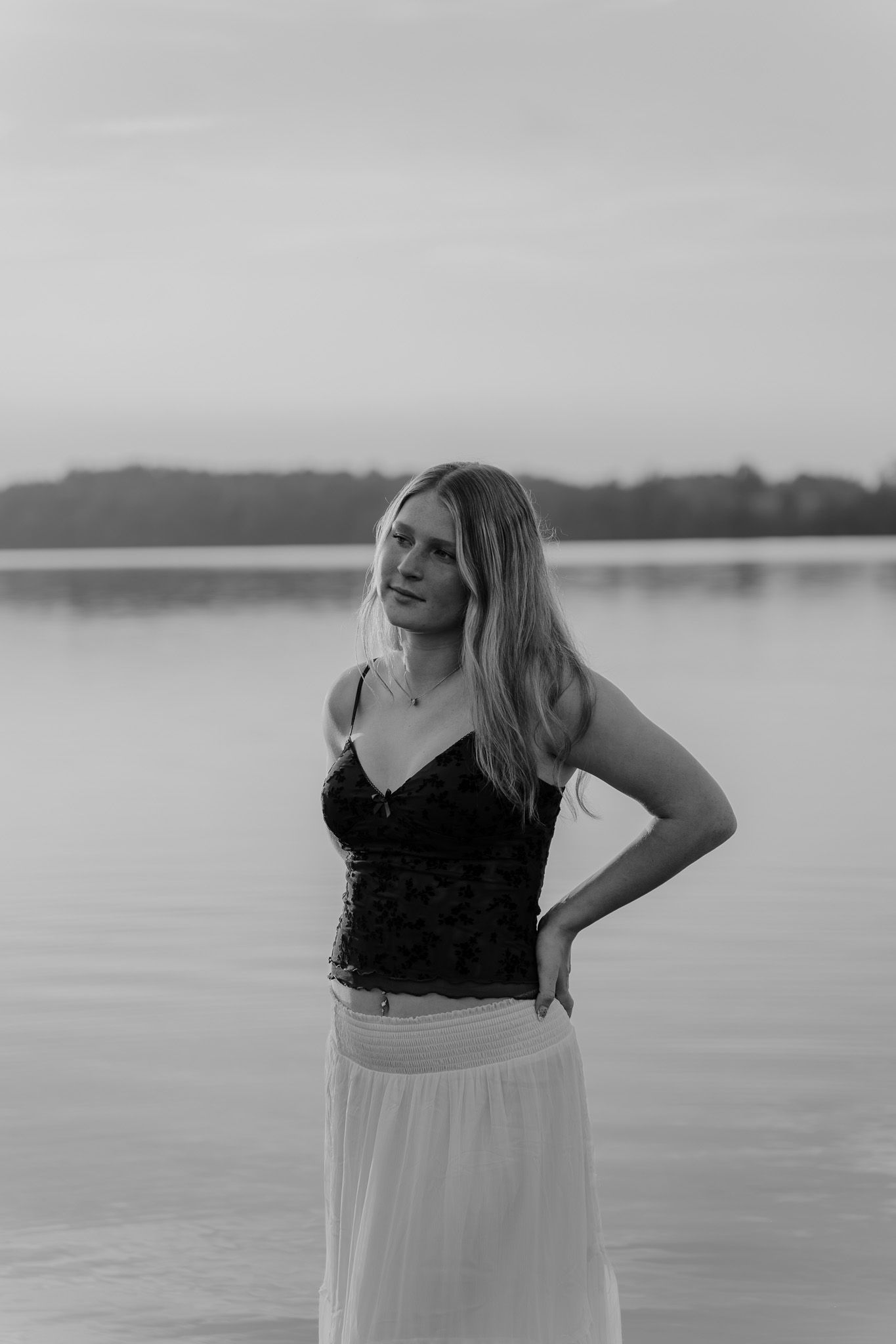 Black and white portrait of blonde woman in lace top and white skirt standing by lake.