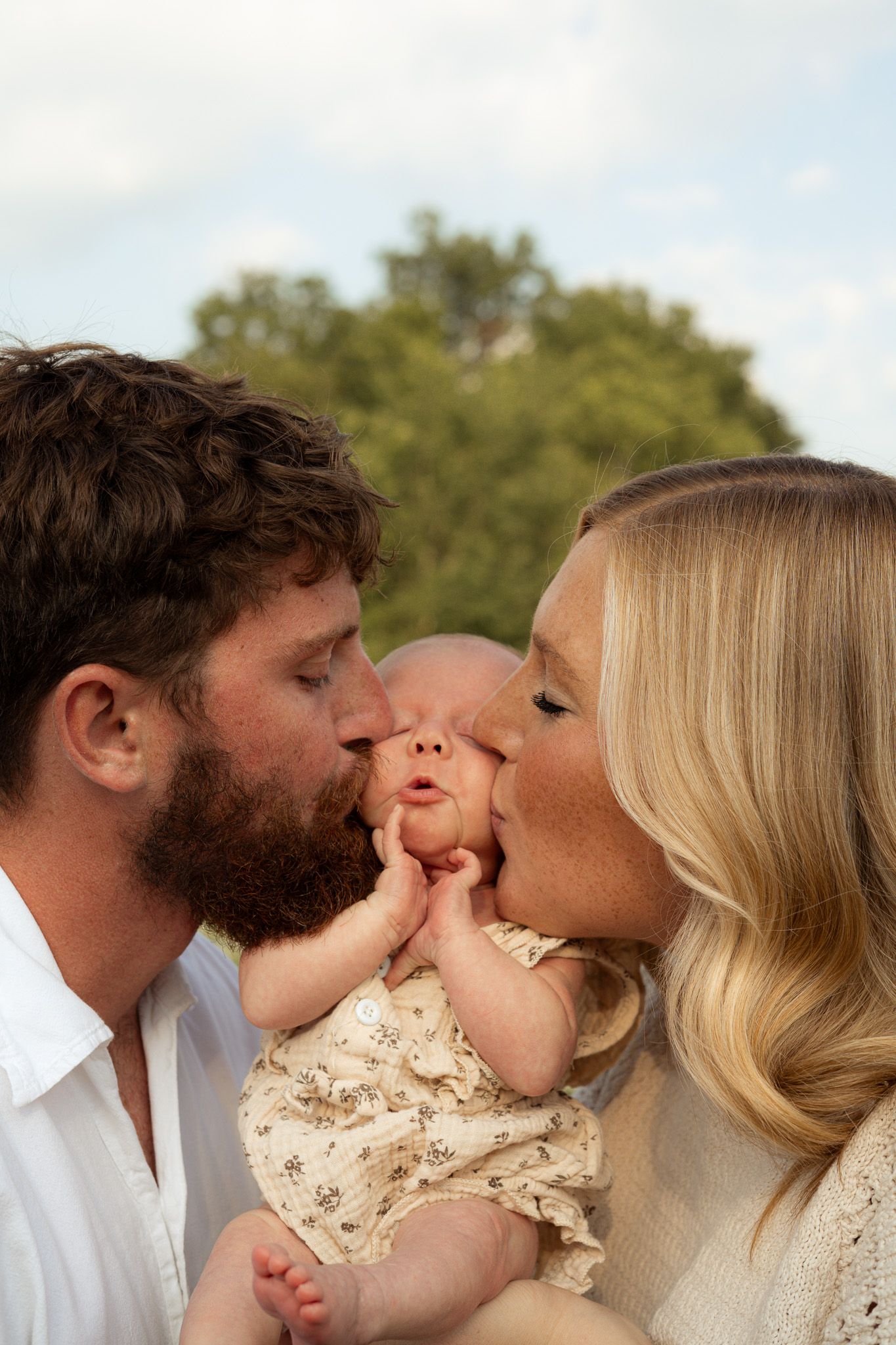 Parents kissing their newborn baby on the cheeks outdoors in a sunny green field.