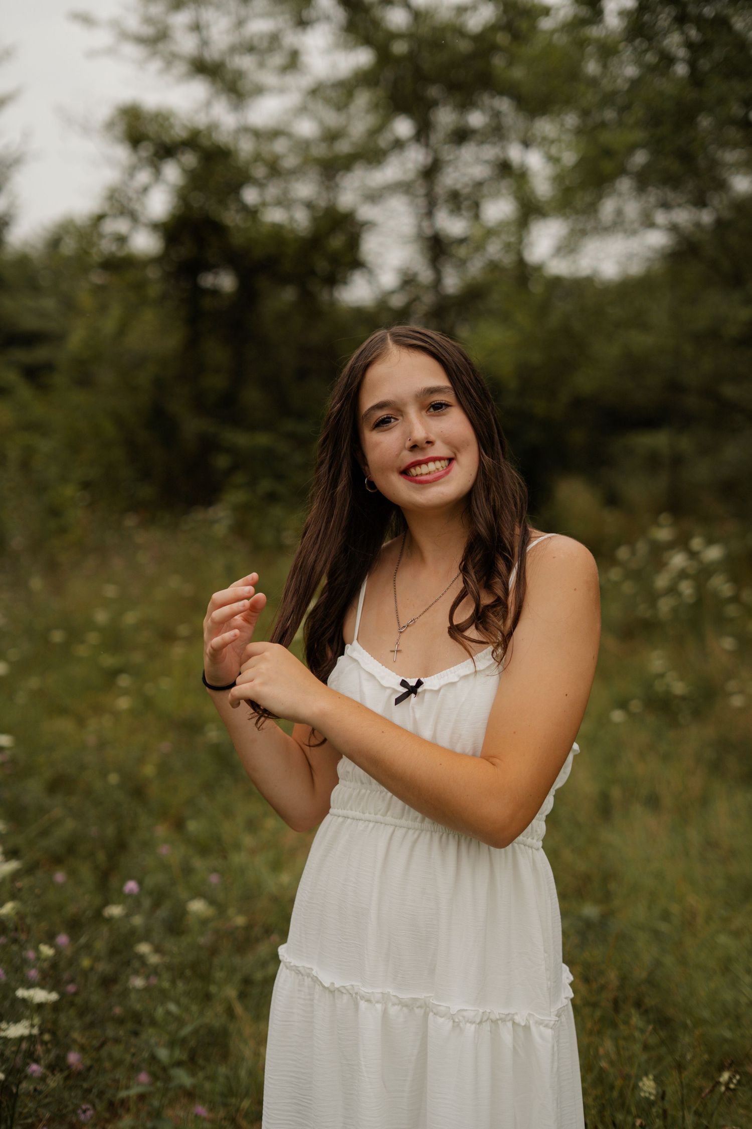 Smiling young woman in white sundress holding a wildflower stem in a lush green field.