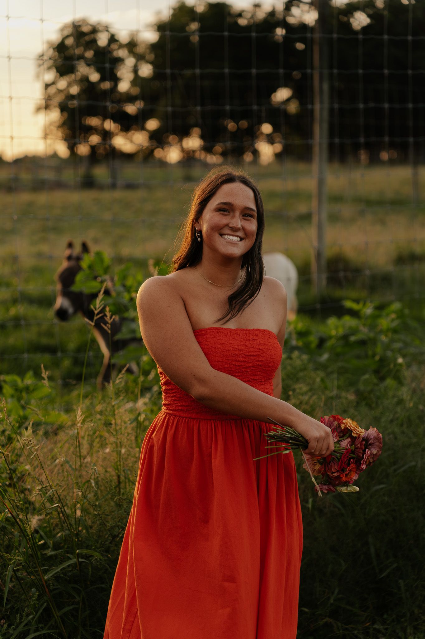 Smiling woman in red dress holding flowers near a fence with horses at sunset.