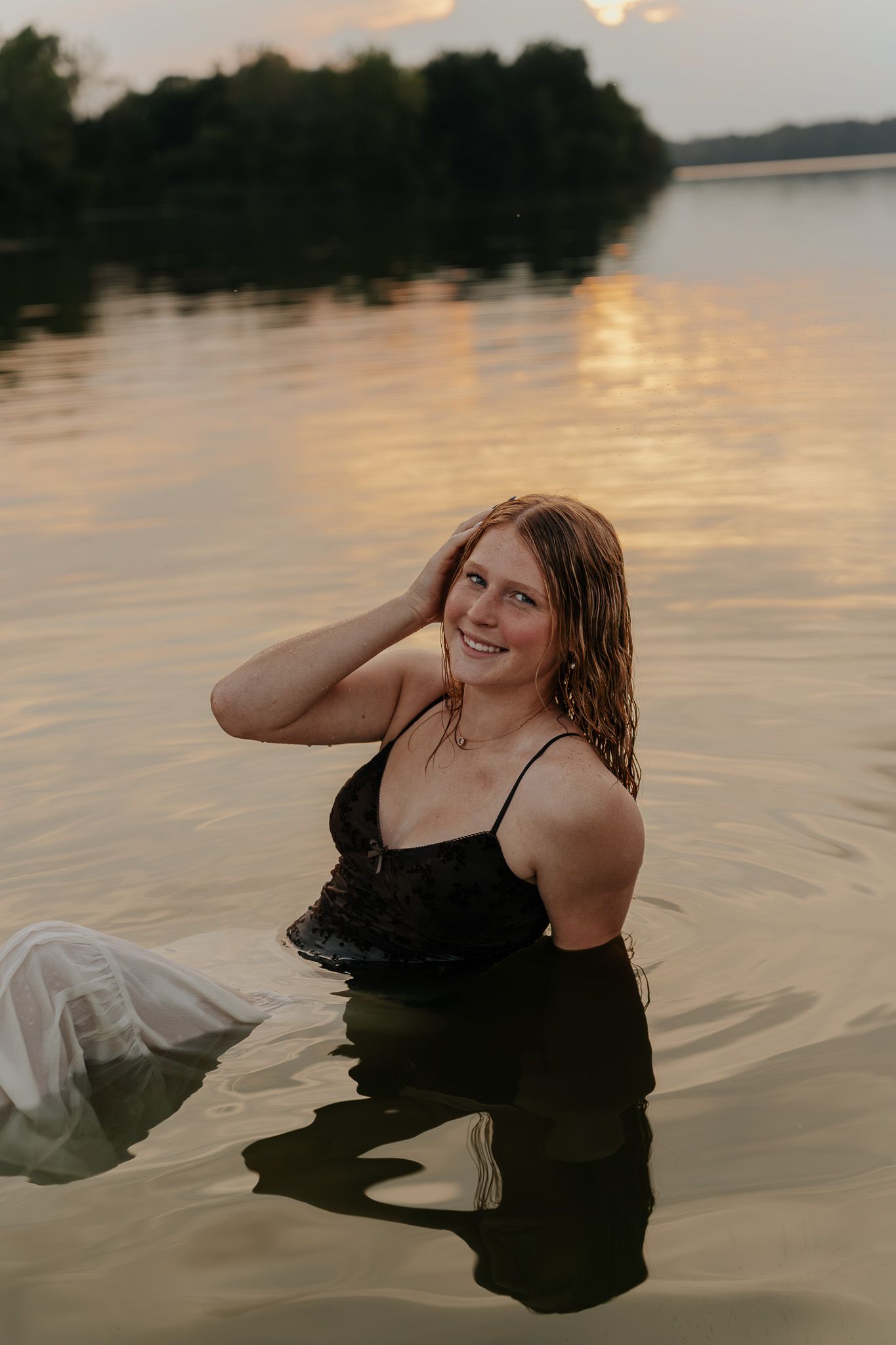 Smiling young woman with wet hair sitting in lake water wearing black top at golden sunset.