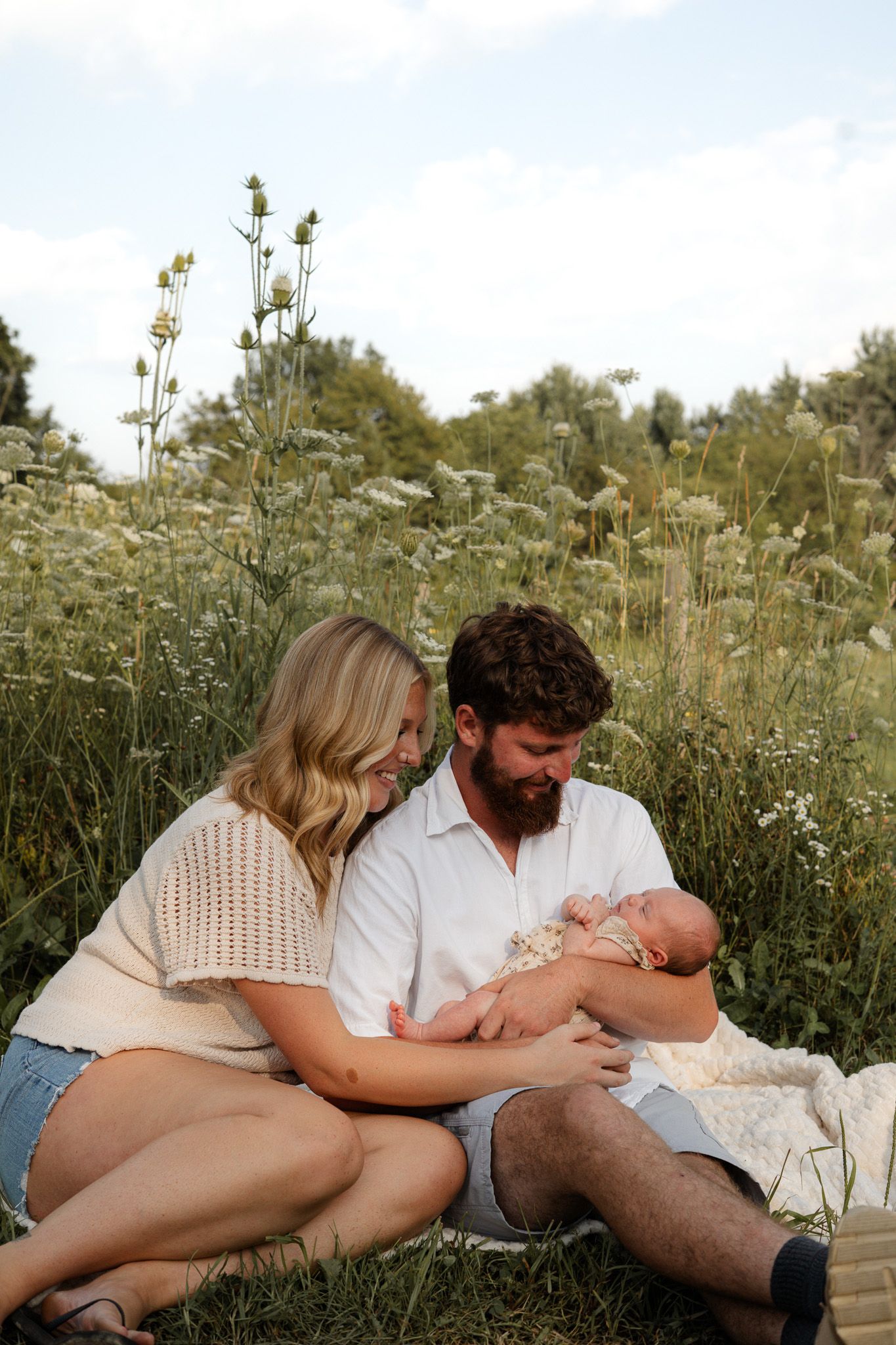 Happy couple sitting in wildflower meadow tenderly holding their newborn baby outdoors.
