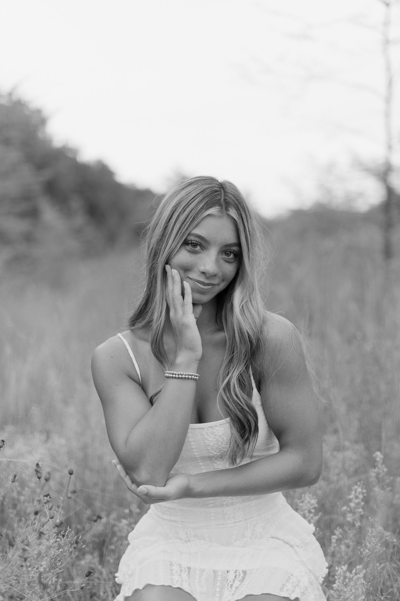 Black and white portrait of young woman in white dress touching face in sunlit meadow.