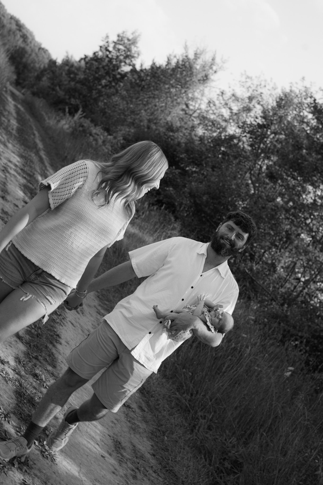 Black and white photo of a young family walking with newborn on a nature path.