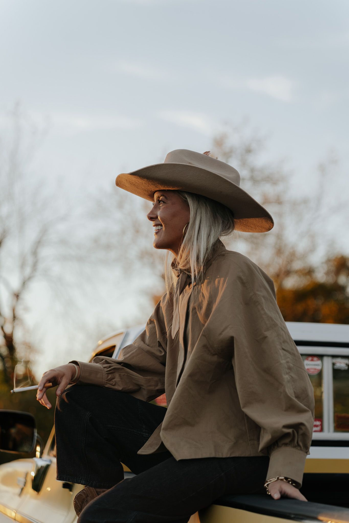Woman in wide-brim hat and tan jacket sits on vintage yellow truck at golden hour.