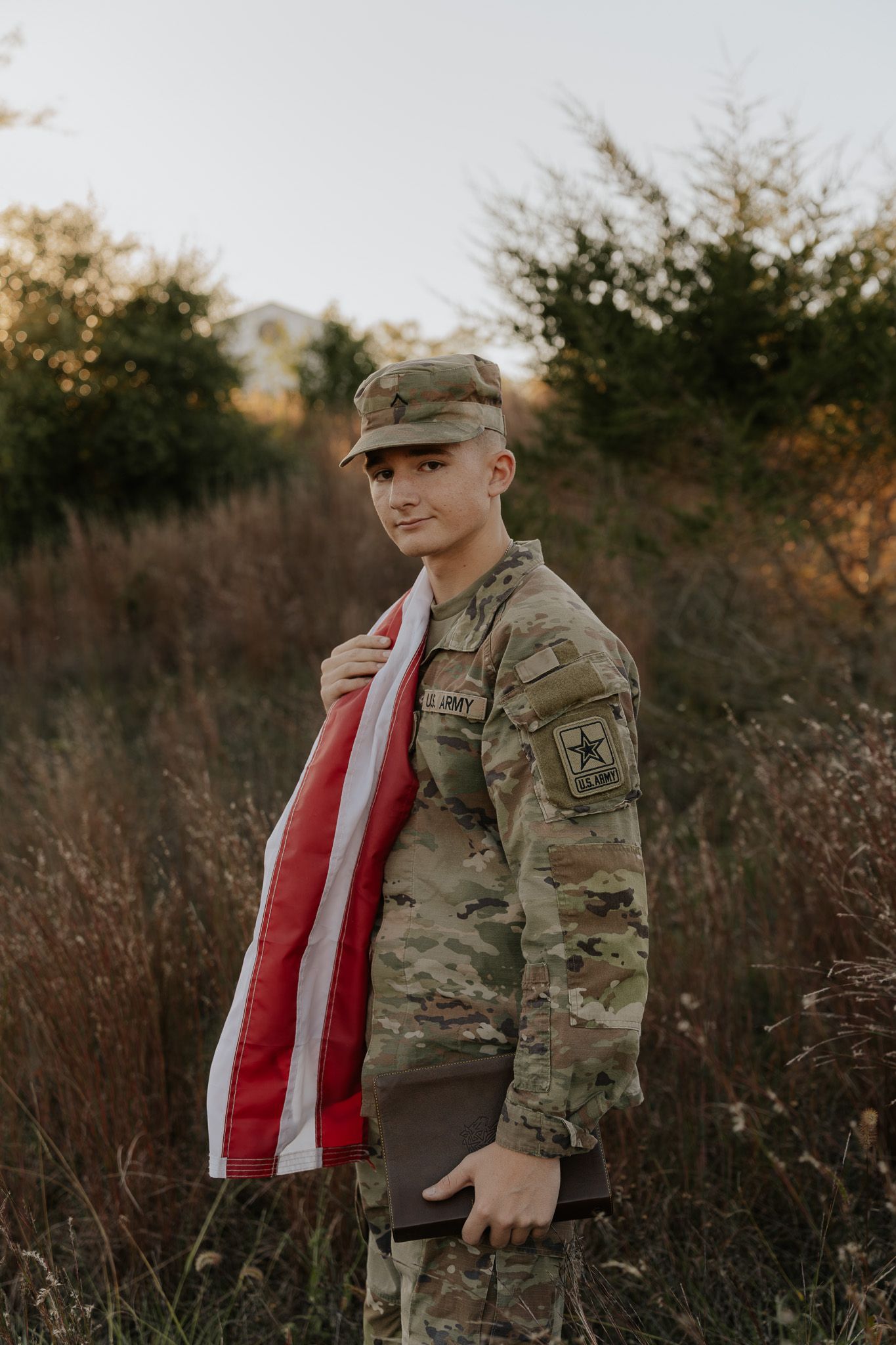 Young US Army soldier in camouflage uniform holding American flag and Bible in a field at golden hour.