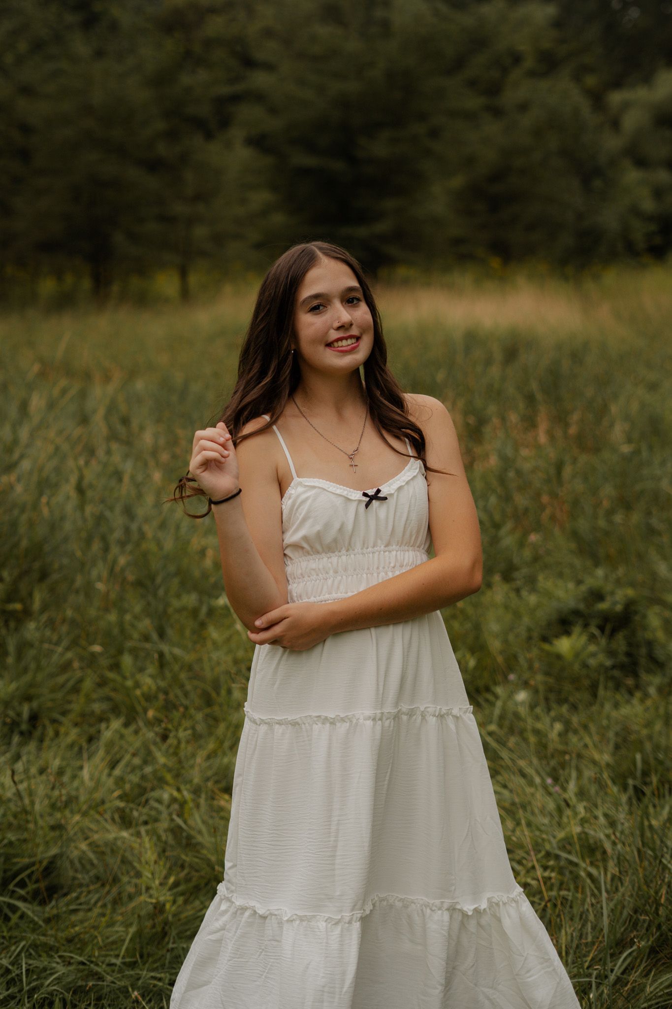 Brunette teen in white tiered sundress standing in green meadow with trees behind.