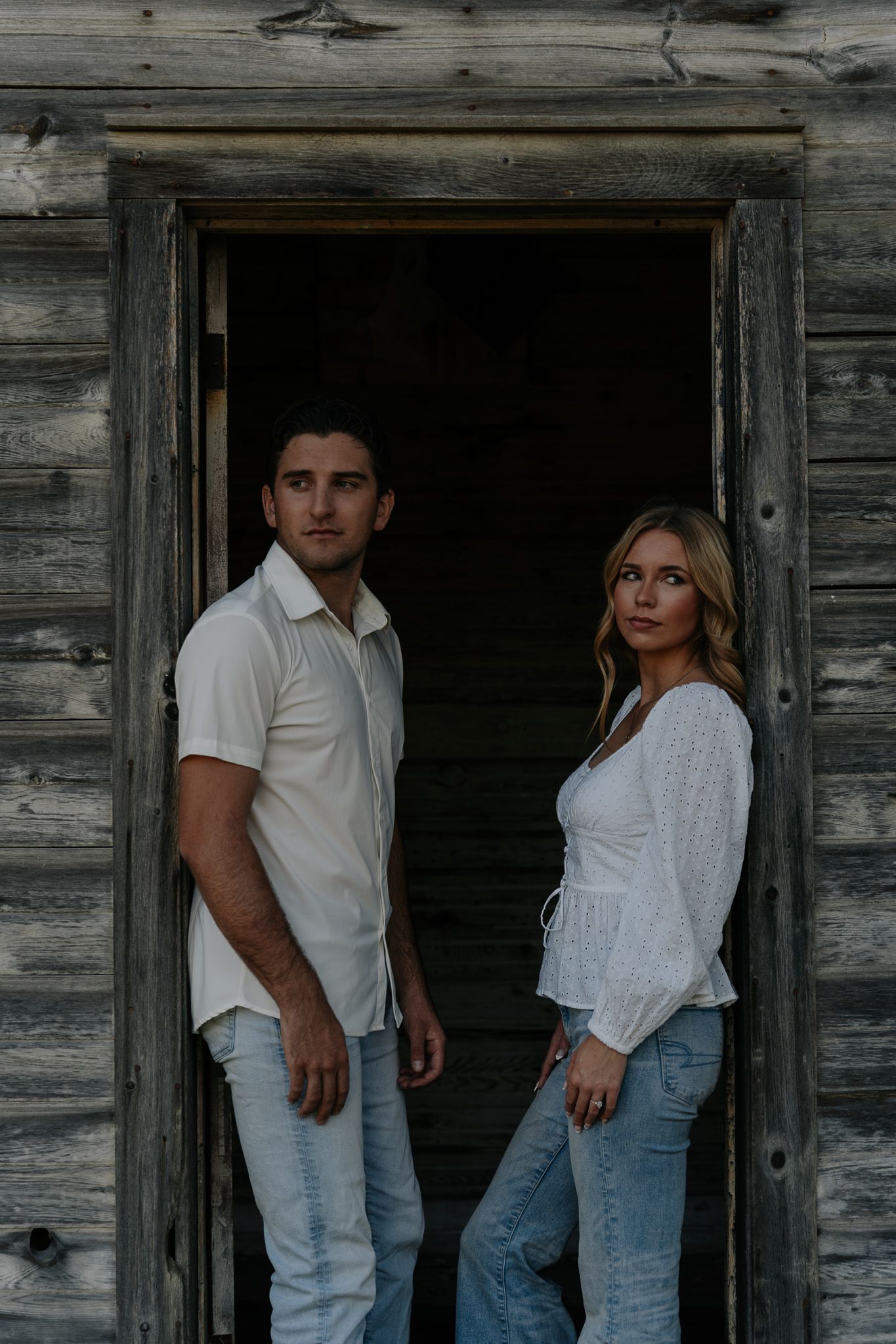 Couple posing in a rustic wooden doorway wearing coordinating white tops and blue jeans.