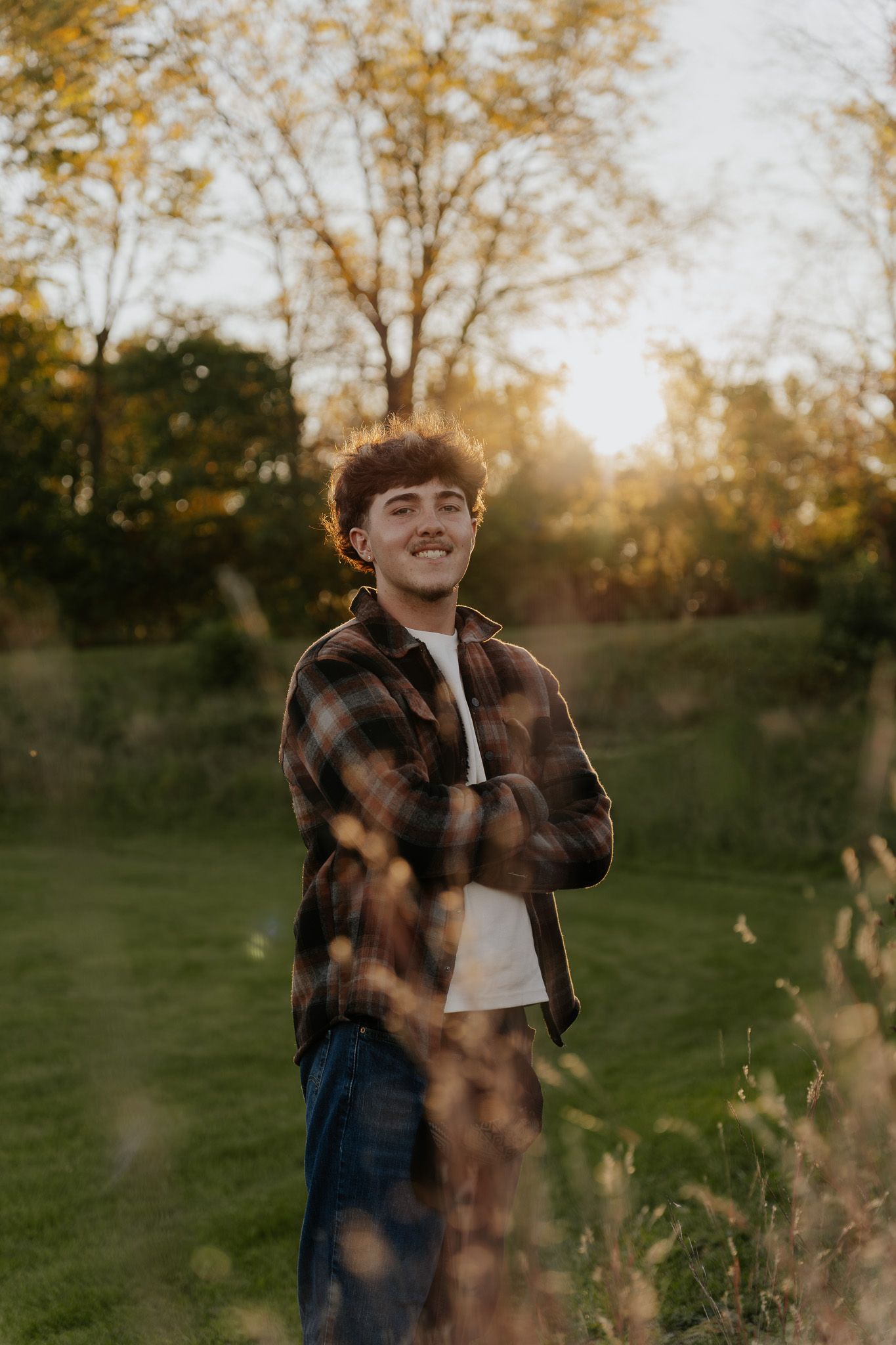 Young man in plaid shirt smiling with arms crossed outdoors at golden hour sunset.