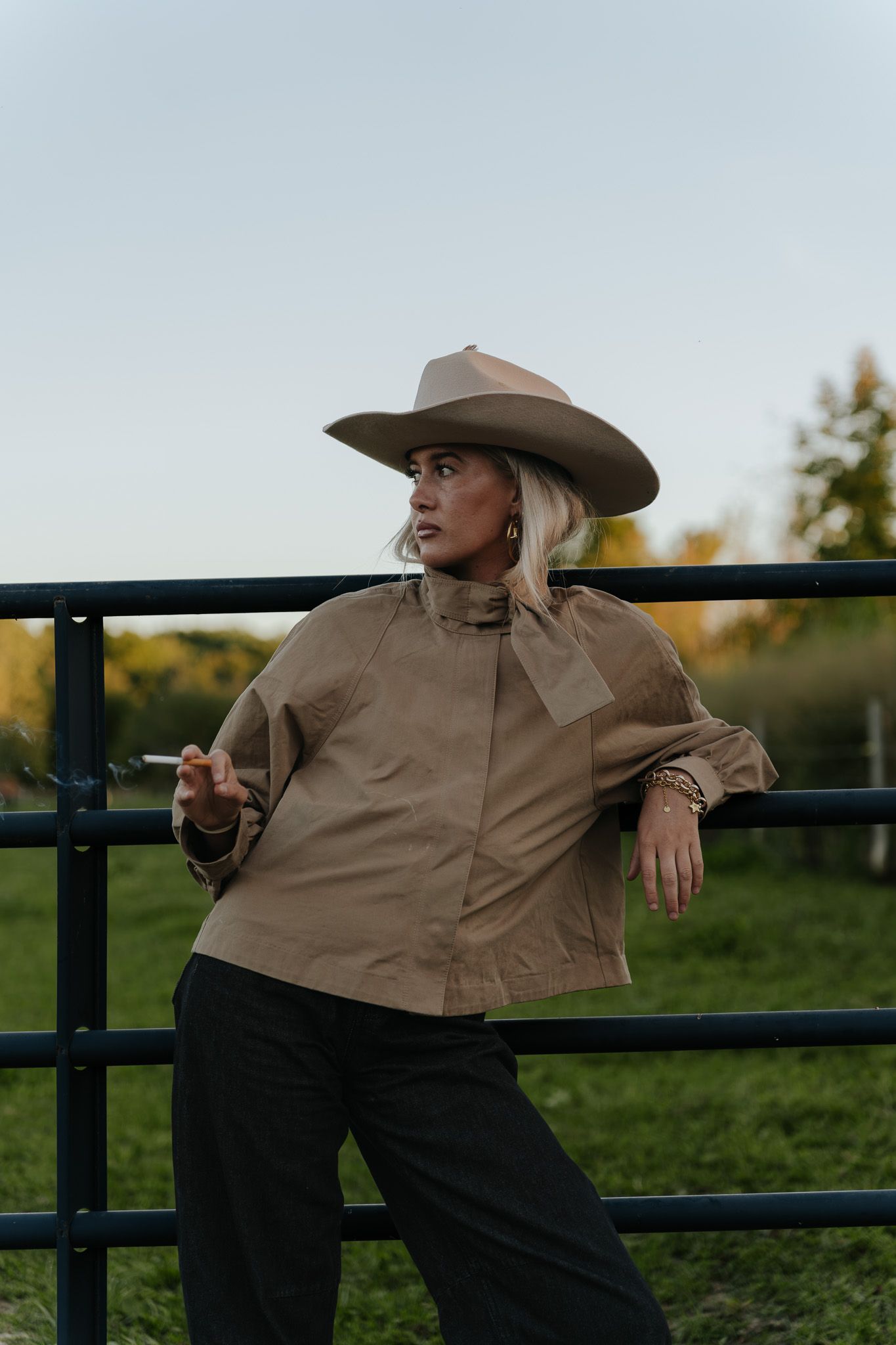 Blonde woman in cowboy hat leaning on a metal gate holding a cigarette at dusk.