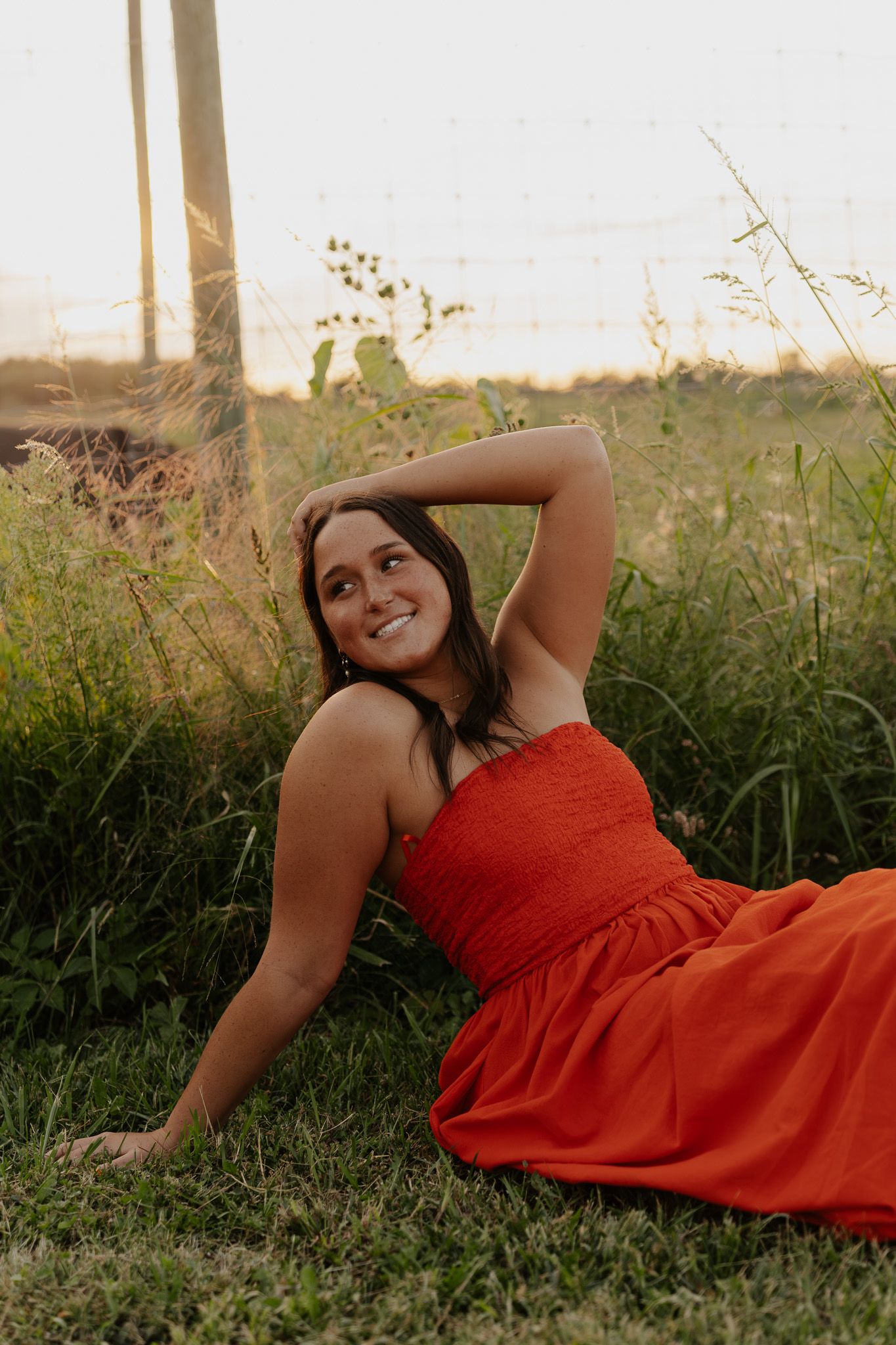 Young woman in red dress sitting in grass smiling at golden hour near a fence.