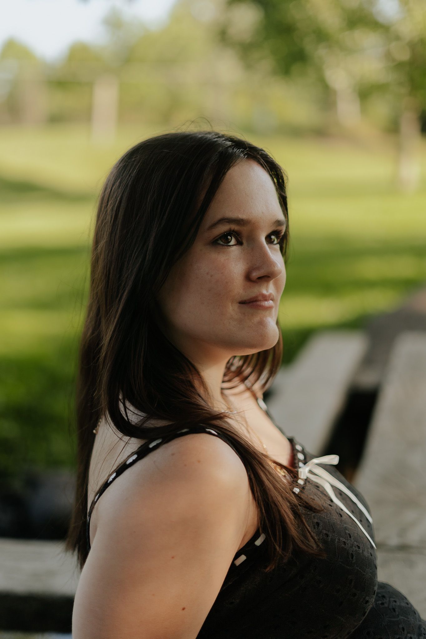 Young brunette woman in black top seated outdoors in sunny park for senior portrait session.