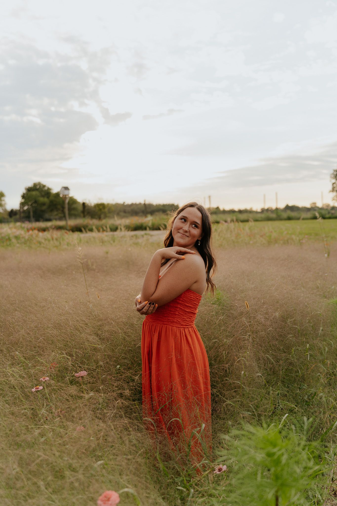 A smiling woman in a red dress stands in a golden field, hugging herself, with a cloudy sky backdrop.