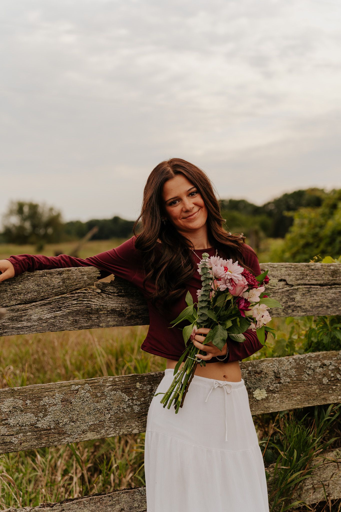 Smiling brunette woman holding pink floral bouquet leans against weathered wooden fence in a lush green meadow.