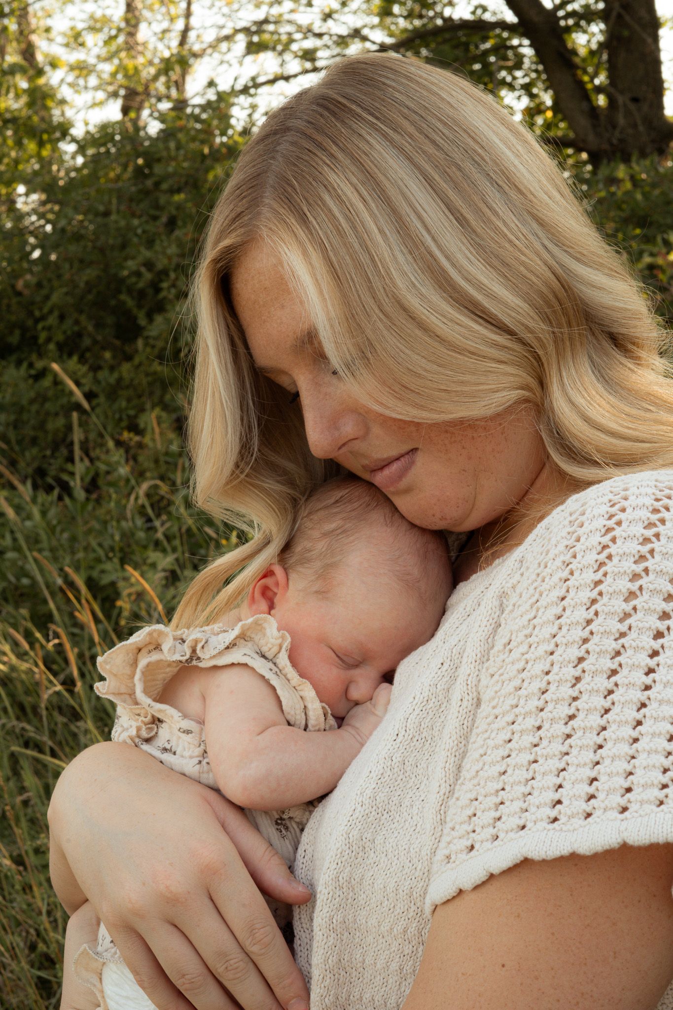 Blonde mother tenderly holding sleeping newborn baby close to her chest in golden outdoor light.