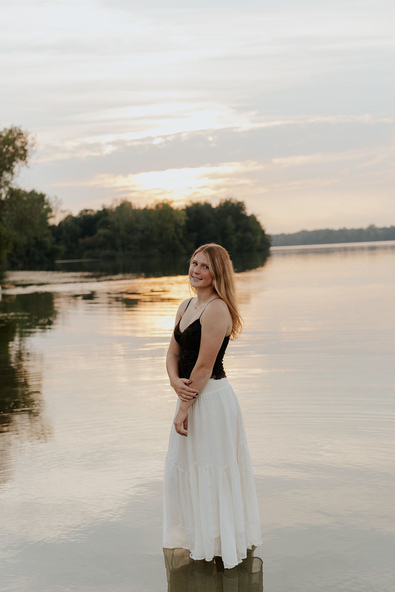 Smiling blonde woman in black top and white skirt standing in calm lake at golden sunset.