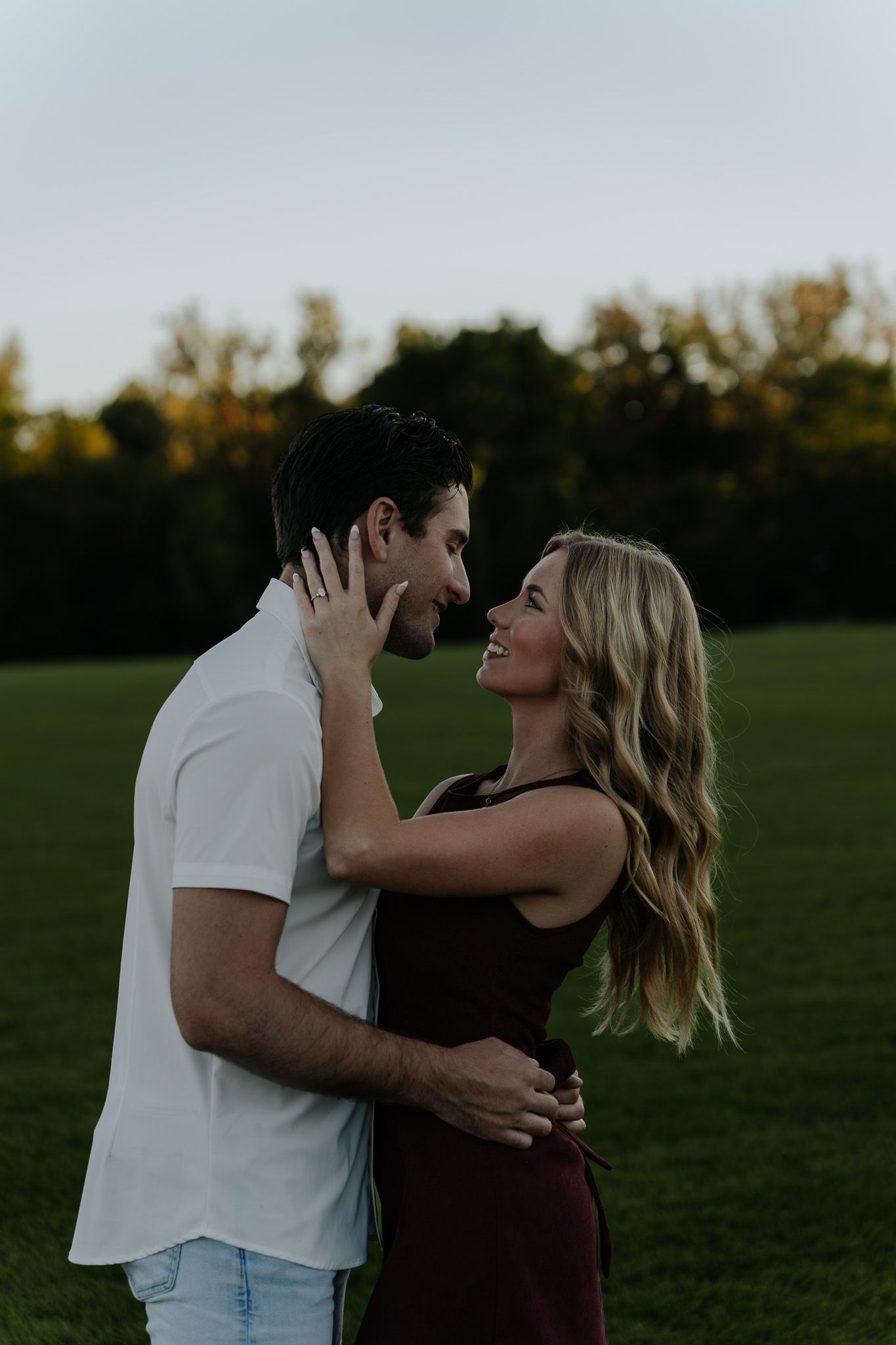 Couple embracing face-to-face in a green field at dusk, sharing a romantic moment.