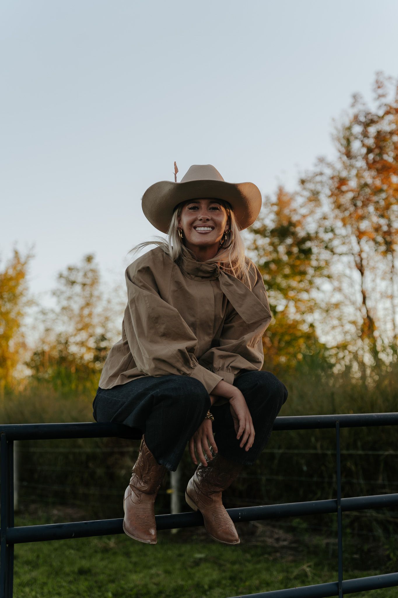 Smiling woman in cowboy hat, tan jacket, and boots sitting on a fence with autumn trees in background.