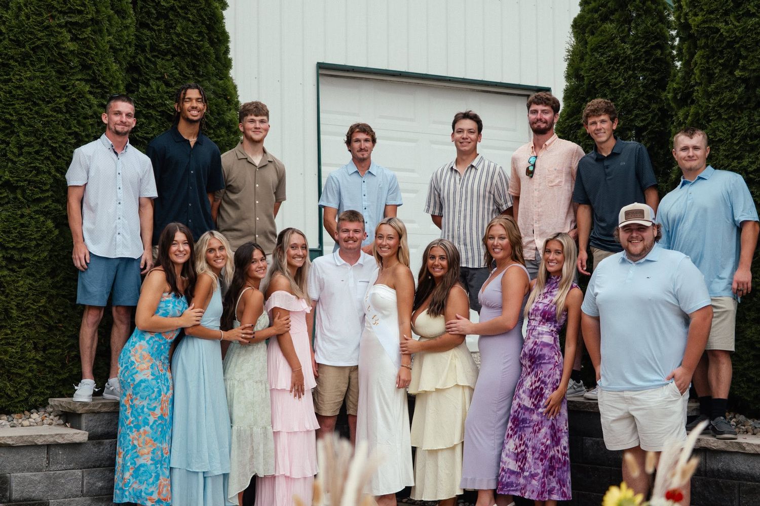 Large group of young adults in formal attire pose together outside a white garage for a celebration.