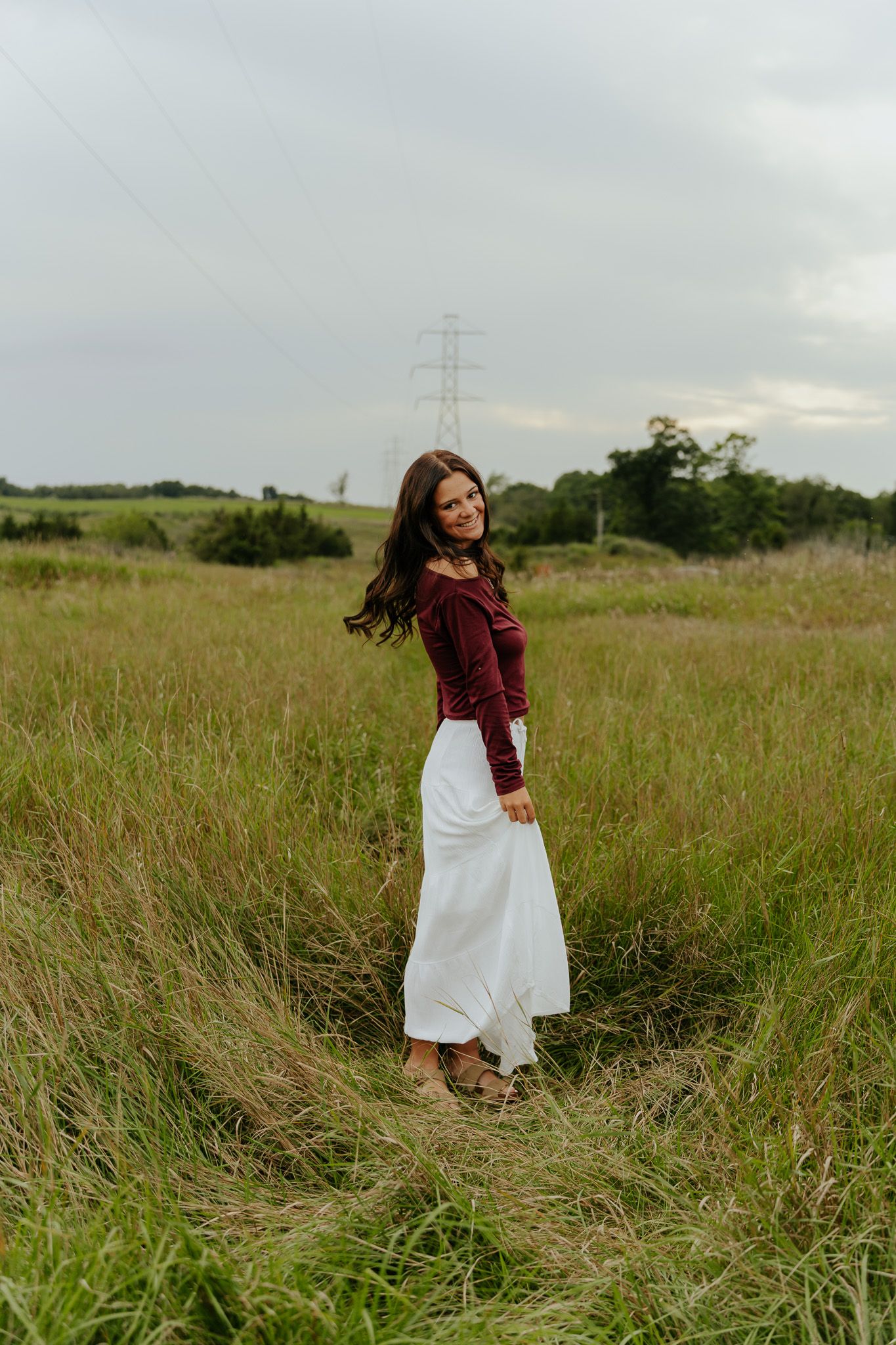 Brunette woman in burgundy top and white skirt posing in tall grass field under cloudy sky.