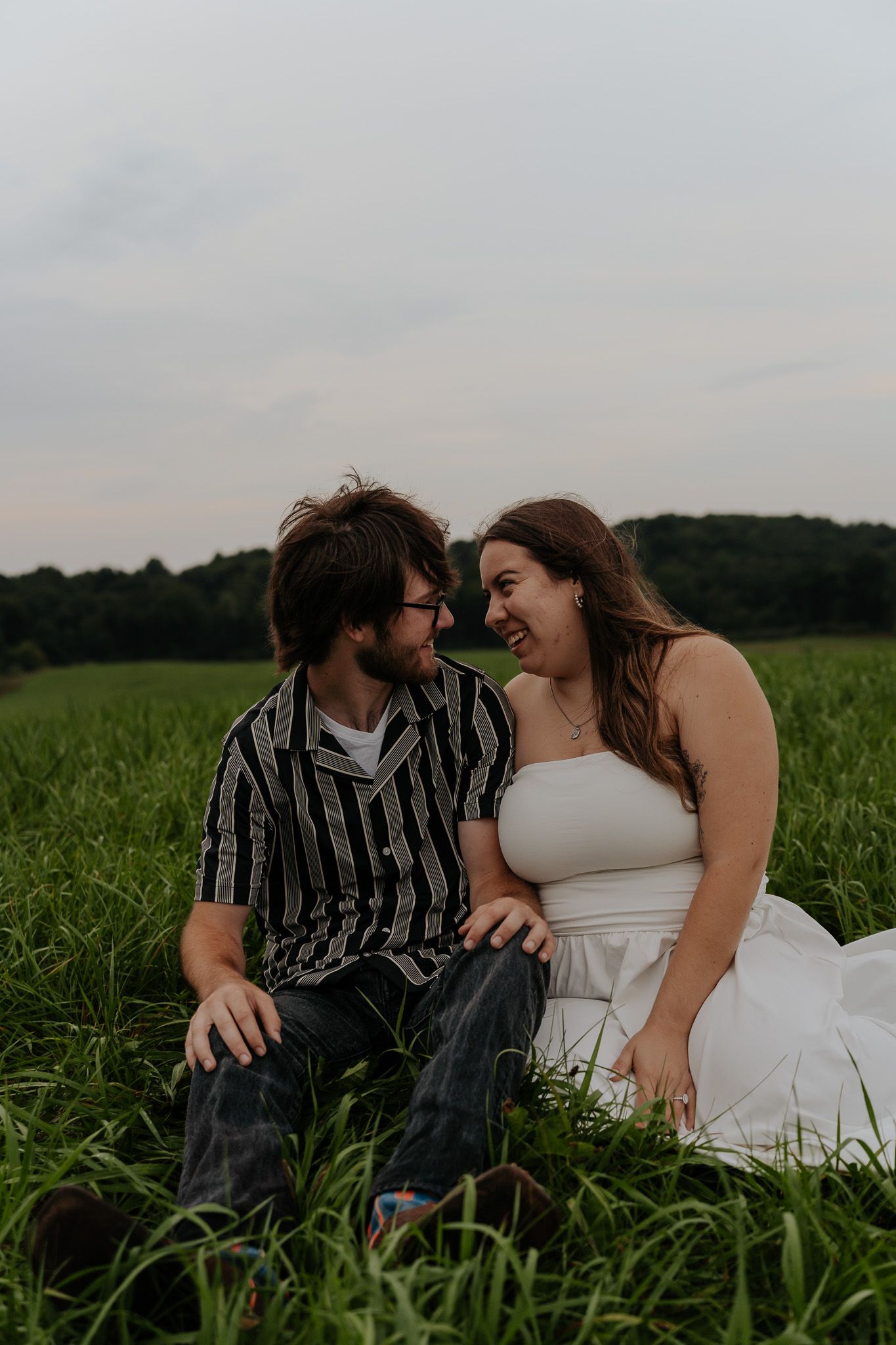 Couple sitting in green field laughing together, woman in white dress and man in striped shirt during engagement session.