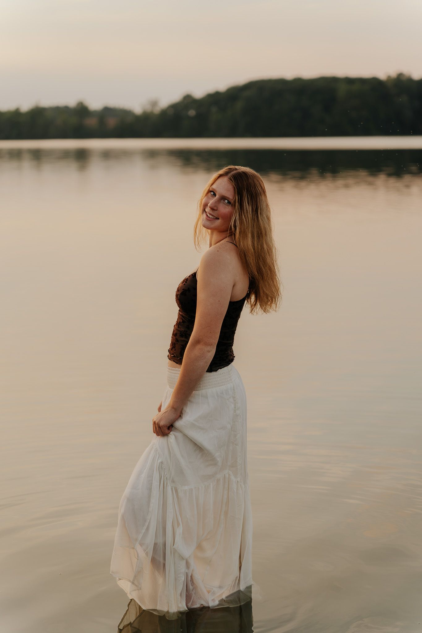 Blonde woman in black top and white skirt smiling over shoulder while standing in lake at dusk.