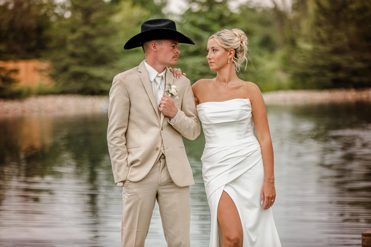 Bride in white strapless gown and groom in tan suit with black cowboy hat pose by a serene lake.
