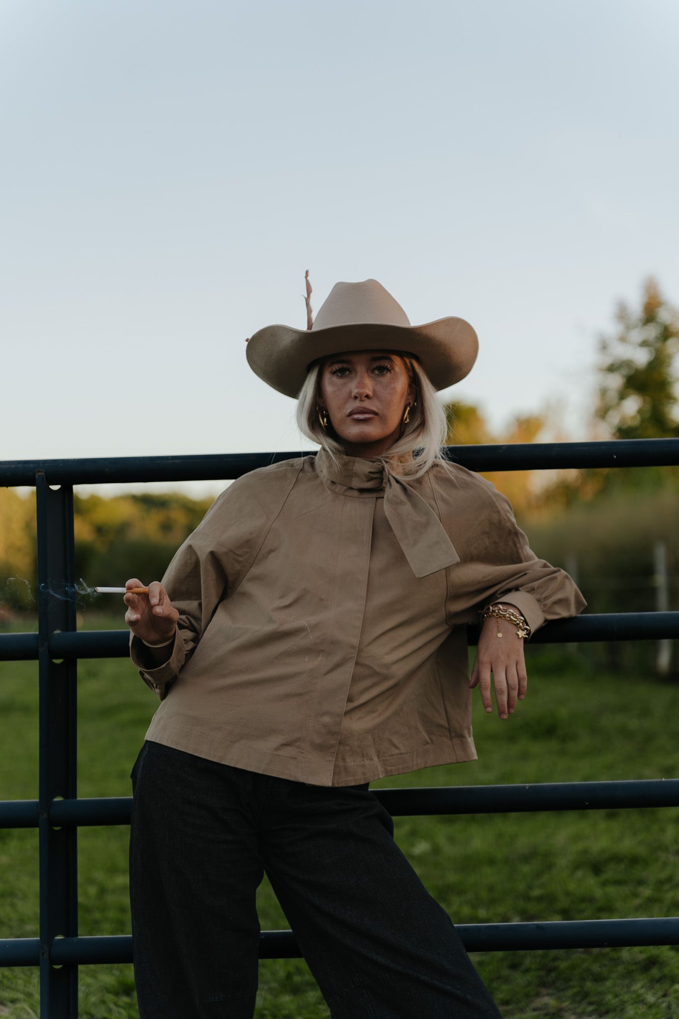 Woman in cowboy hat and tan shirt leaning on metal fence in rural field at dusk.