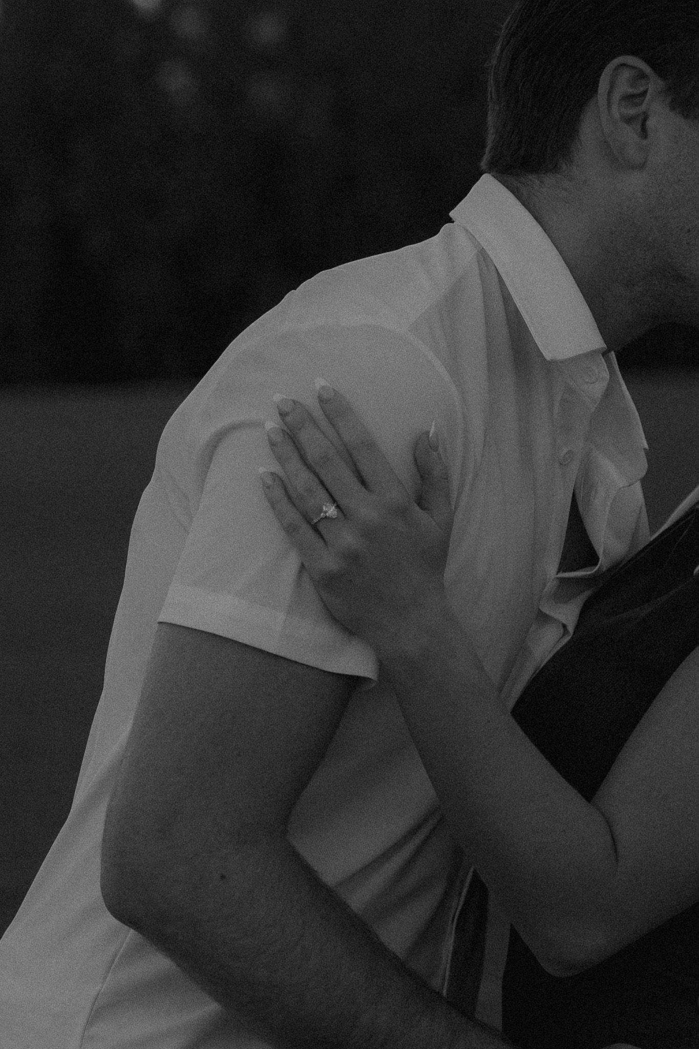 Black and white photo of a couple embracing, woman's hand with engagement ring resting on man's shoulder.