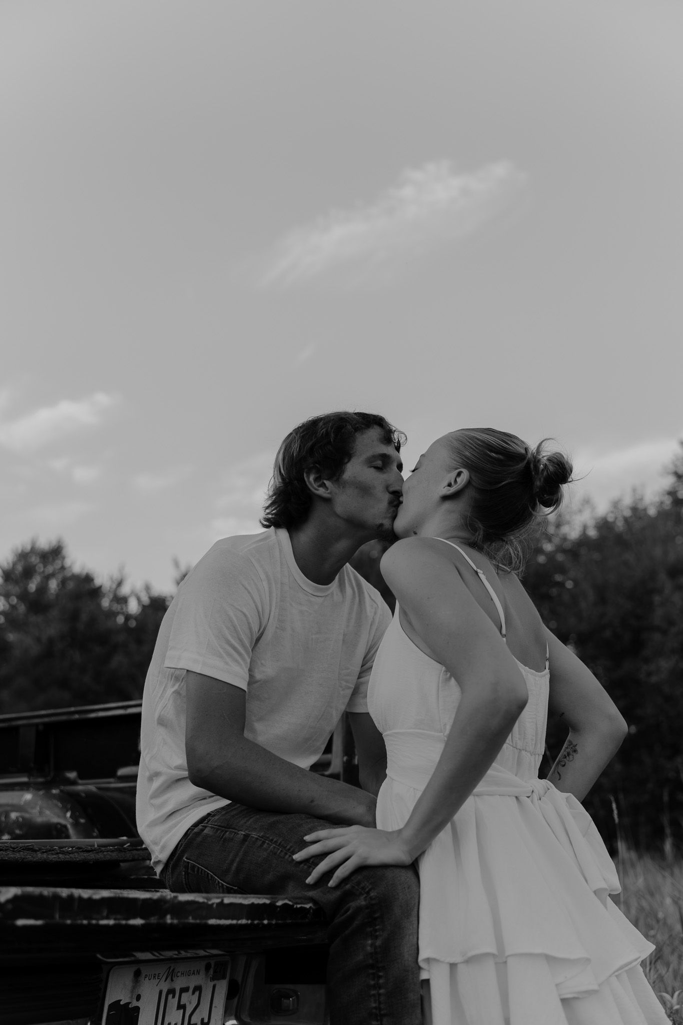 A couple shares a romantic kiss leaning against a vintage truck in a black and white photo.