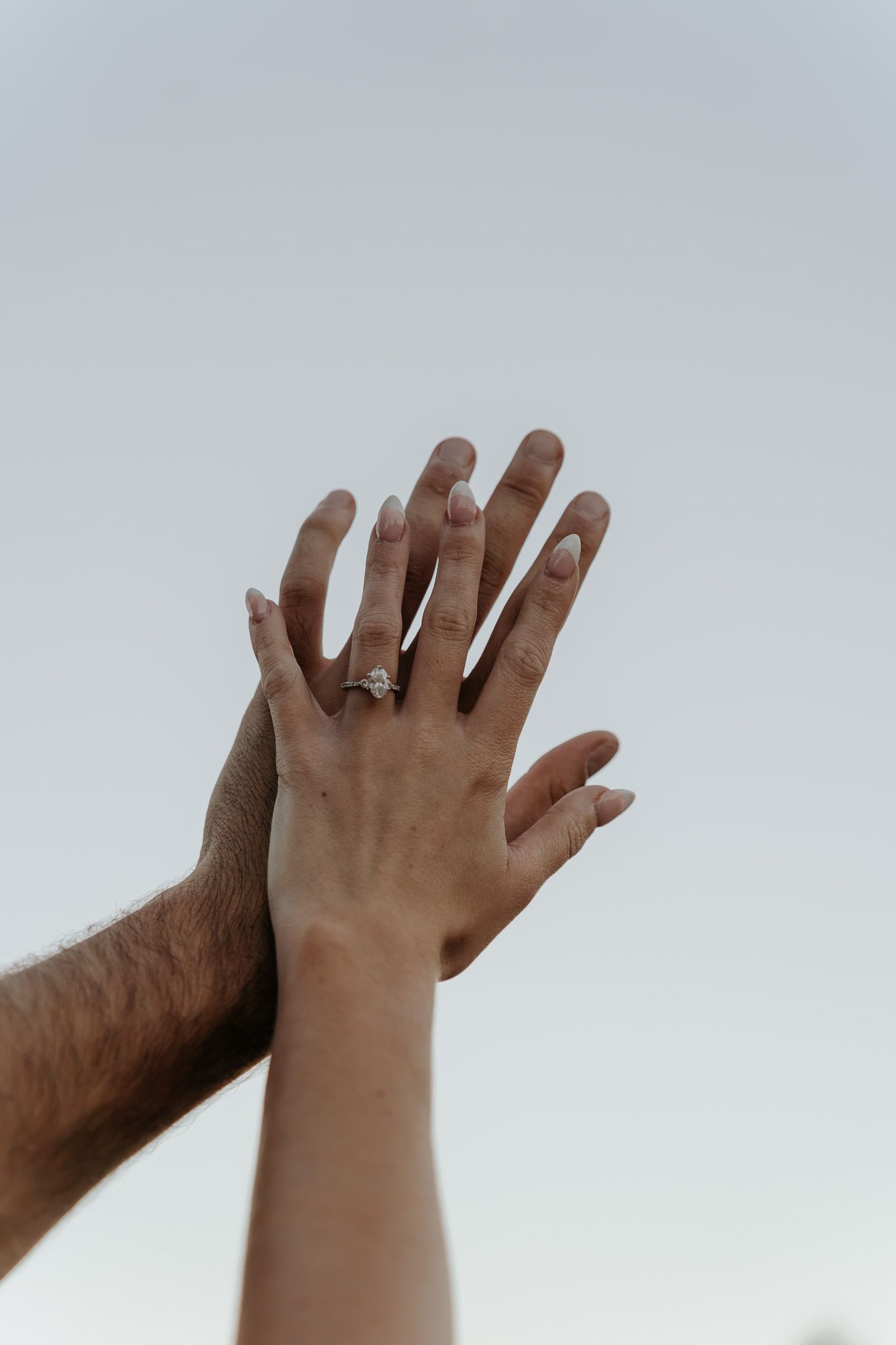 Couple raising their hands together against a bright sky, showcasing an engagement ring.