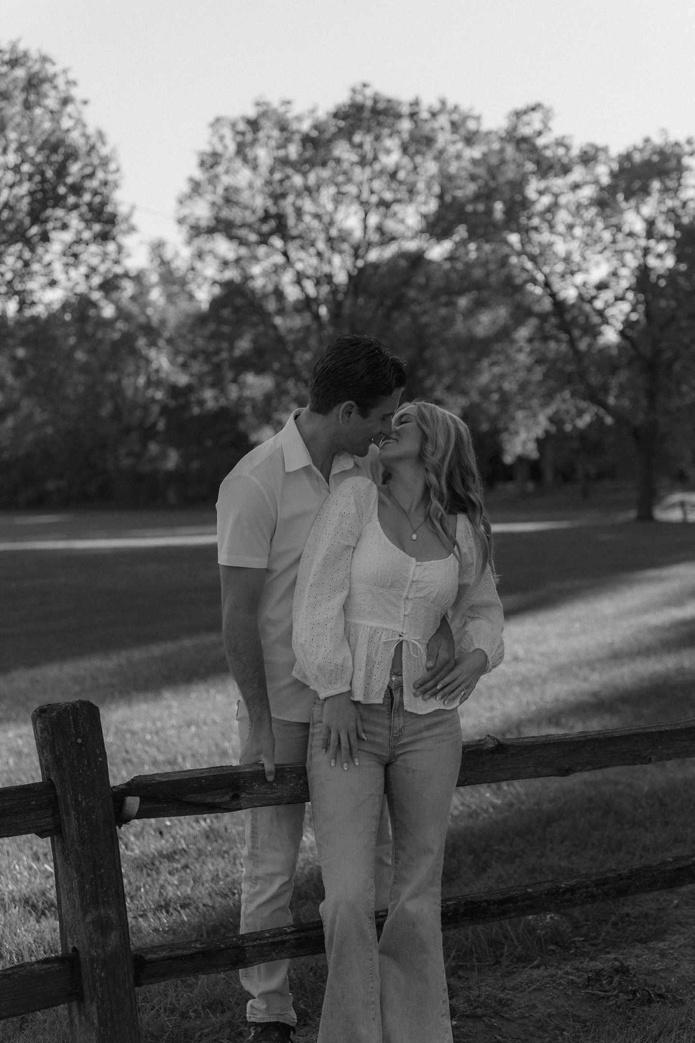 Black and white photo of a couple sharing a kiss near a wooden fence in a lush park setting.