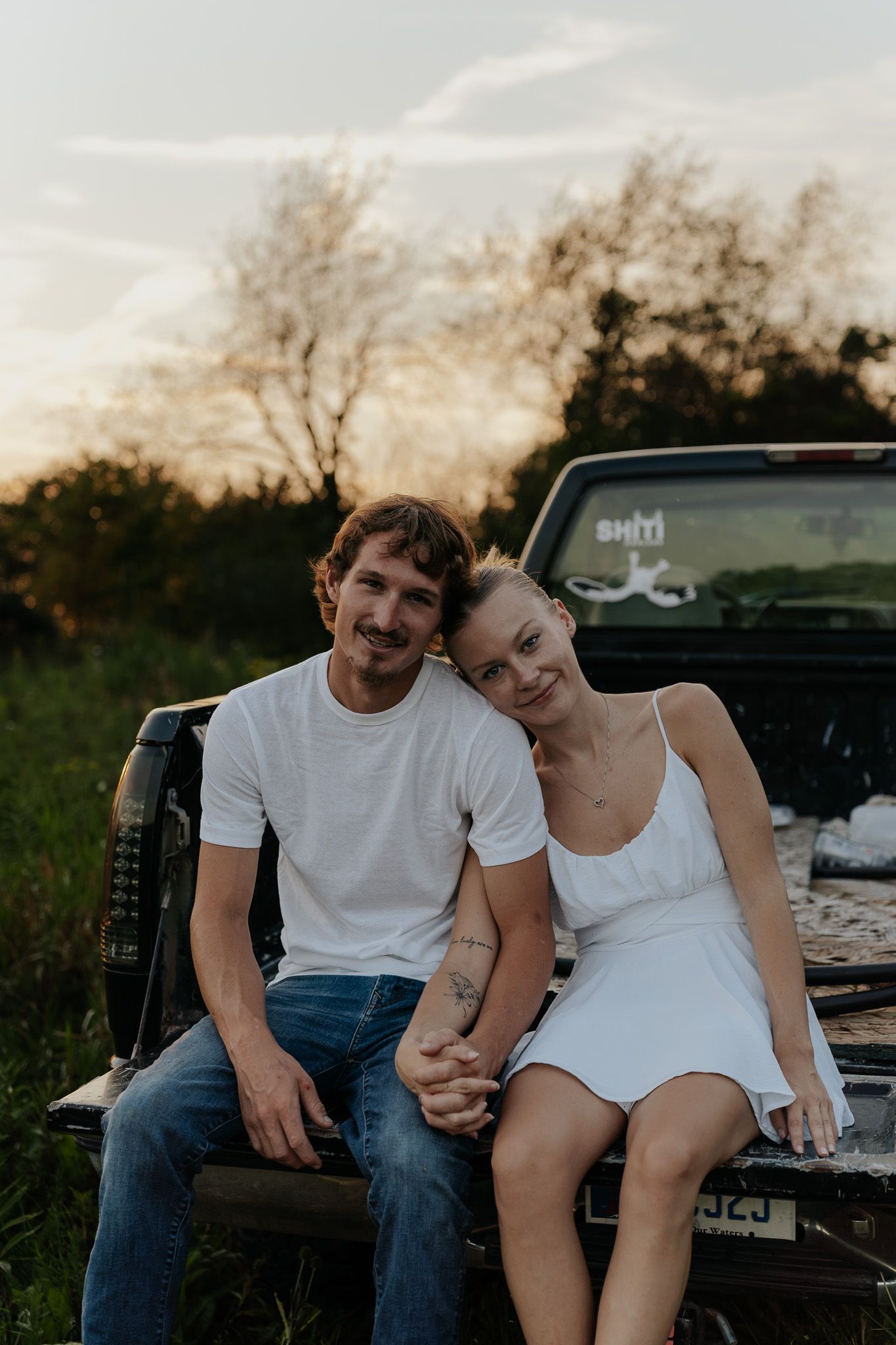 A smiling couple in white outfits sits together on a vintage truck tailgate at sunset.